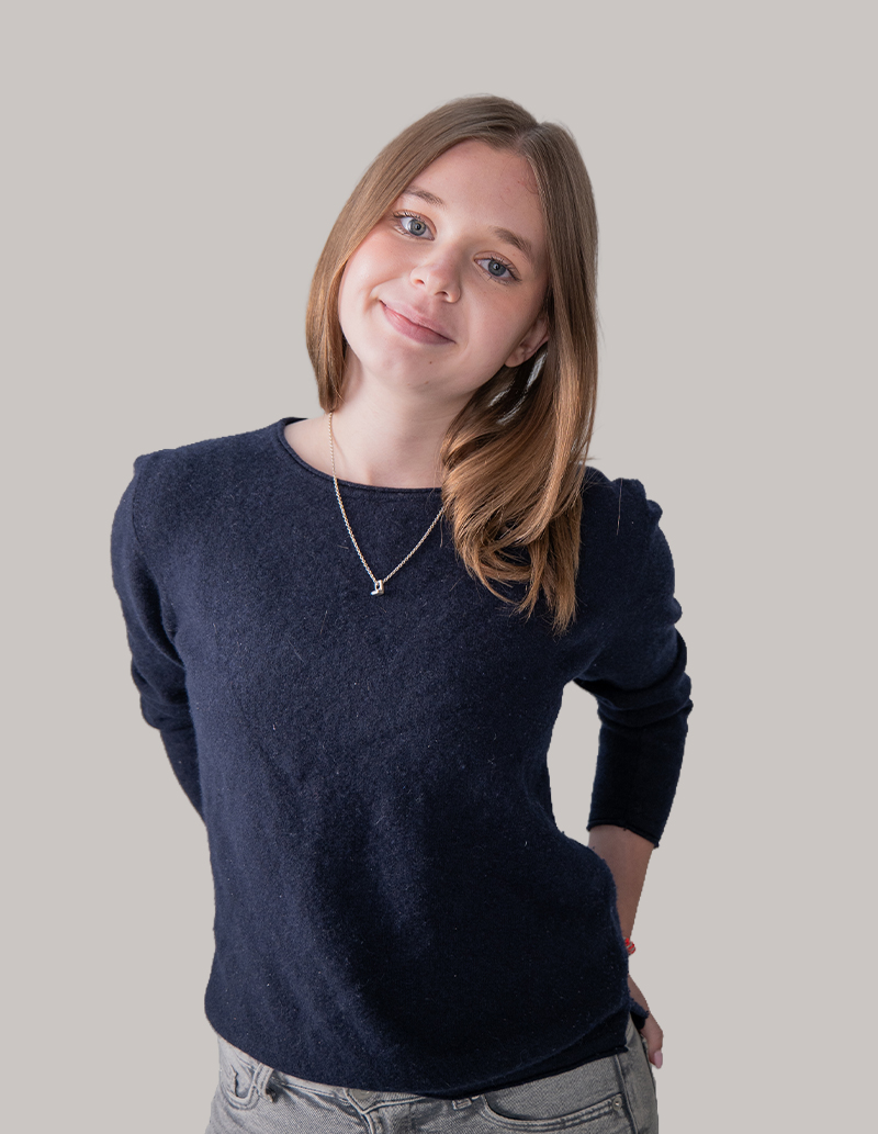 Eine junge Frau mit glattem, hellbraunem Haar steht vor einem schlichten Hintergrund und verkörpert die Professionalität, die man oft im Branding und Webdesign von Agenturen sieht. Sie trägt einen marineblauen Pullover, eine graue Hose und eine Halskette und lächelt sanft, während sie die Hände hinter dem Rücken verschränkt hat.